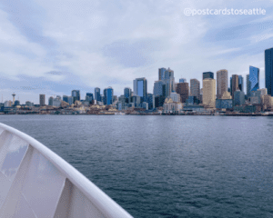 Bow of boat in foreground, blue water in midground, and tall, multicolored buildings in background