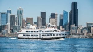 Large boat with passengers and skyline behind with blue sky and waves in foreground